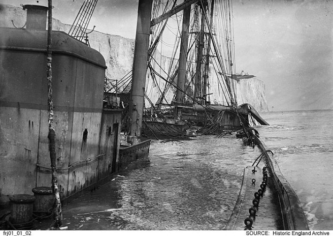 B&W photo of shipwreck showing amidships structure and masts to left, deck awash with water from the sea to the right, cliffs in background.