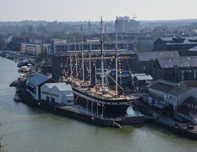 Modern colour photograph seen looking down from a nearby window over the SS Great Britain displayed in dock and 'dressed overall' with flags flying between her six masts. and the city beyond. The river is seen to left and foreground.