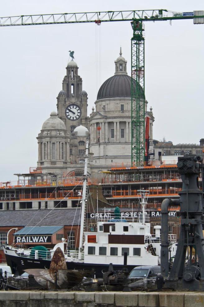 Modern colour photograph of Liverpool: edge of dock in foreground with propeller to left and steam crane to right, with the black & white hull of the ship just beyond in the middle ground. The deck lies under the lower roof of the warehouse saying RAILWAY, while the upper structure lies beneath the upper roof structure of the warehouse with the words GREAT WESTERN RAILWAY, creating a visual echo in the composition, also echoed by the building works beyond, and the varied heights and sizes of the Port of Liverpool and Royal Liver Building domes at the top of the image.