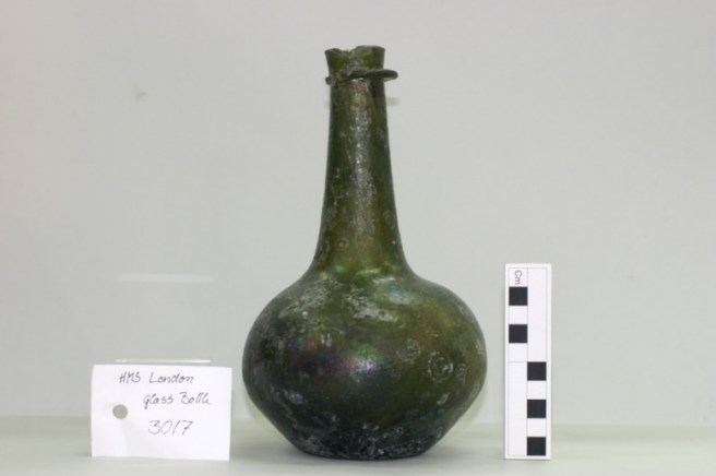 Rounded dark green glass bottle with tag to left and scale rule to right.