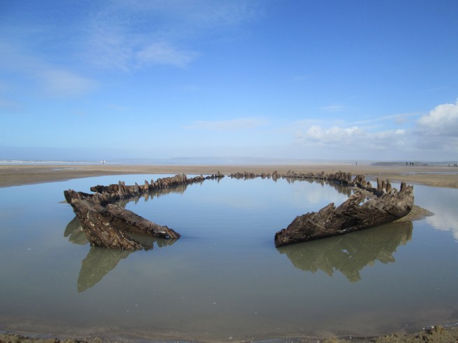 The almost complete outline of a wreck sitting in a pool of water against a backdrop of blue sky and clouds reflected in the water.