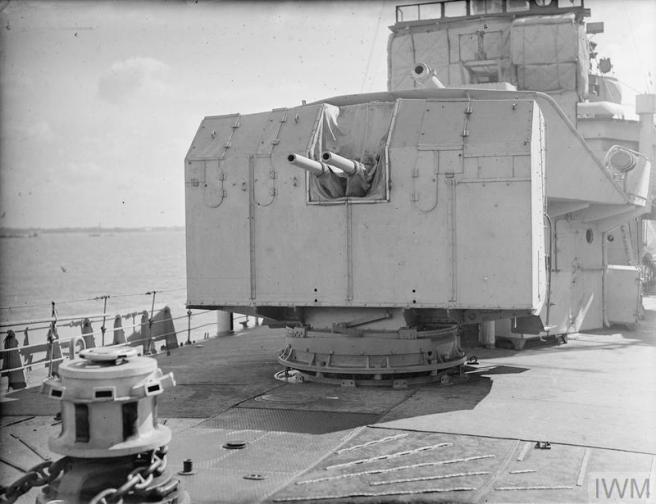 Historic B&W photograph of guns mounted on a swivelling turret with a further gun mounted above on the deck of a ship in harbour. Across the sea in the distance are other ships dotted along the coastline.