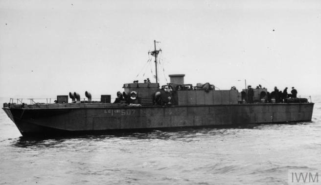 Historic black and white photo of landing craft profiled in port view, showing a long, low craft and men clustered astern (to right of image)
