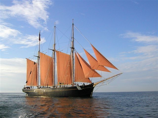 A ship in the centre of the image sits against a blue sky, the sea occupying the bottom third of the image. The ship has three masts, with three square red sails spread, and four triangular sails between the foremast and bowsprit, which faces to the right of the image.