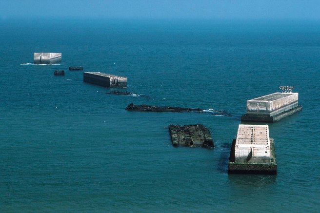 Modern colour photograph of 8 sections of Mulberry Harbour in a bright blue sea, in various states of decay and angles from each other, no longer a coherent harbour assemblage.