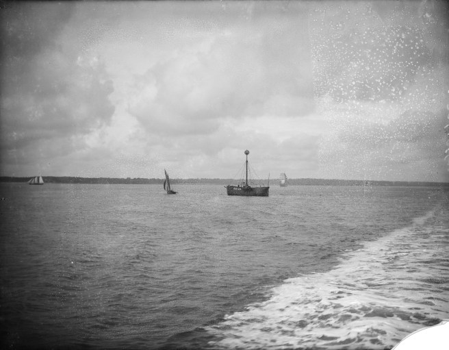 Historic black and white photograph looking towards a lightship at sea, identified by its mast and the name 'CALS. . . ' visible in large white letters to the right of the hull, and a small yacht to the left. A tidal wash is visible to right of the image, which is compromised by a broken right-hand corner and other damage across the upper sky visible in the original glass plate negative.