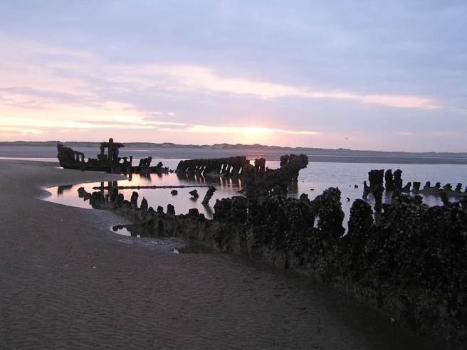 View of shipwreck on sands at sunset, showing skeleton of vessel.