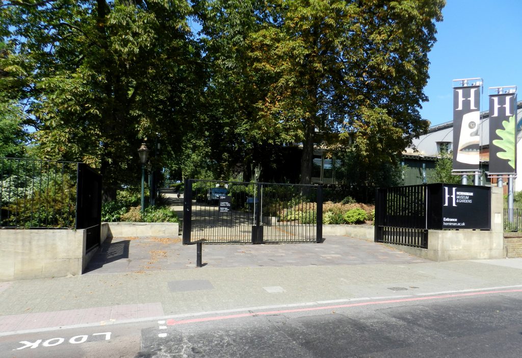 A picture taken from a road towards some black gates, which have two open sections to either side and a central closed panel. A sign to the right says Horniman Museum and Gardens entrance horniman.ac.uk. There are gree trees and a path leading lighly upward in a straight line. There is an old fashion lamp just inside the gates, and some low buildings to the right