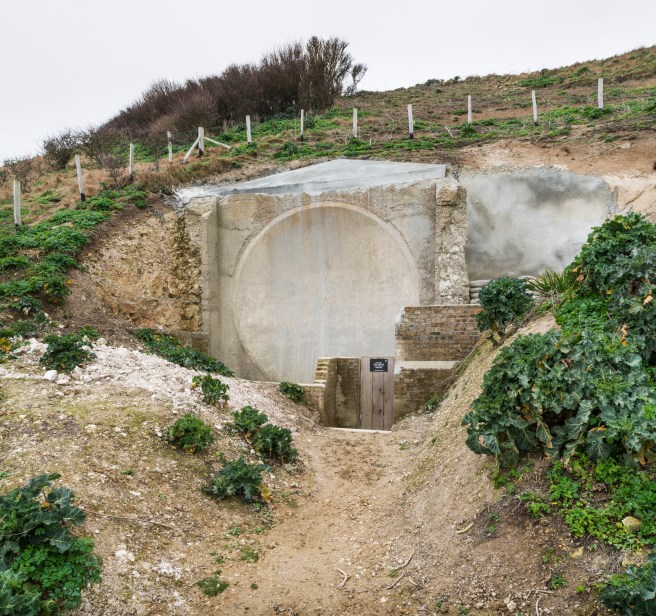 Modern colour photograph of a circular structure cut into a chalk cliff at the centre of the image, surrounded by green vegetation.