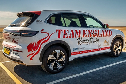 Branded Team England and Sport England electric taxi parked by a wide sandy beach on a sunny day under a clear blue sky.