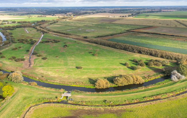 Arial shot of fields and trees, a river runs through the bottom of the photo and the skyline is in the distance. 