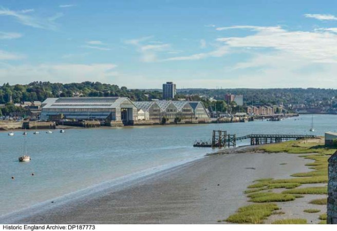 Colour photograph of blue river running diagonally through image from bottom left corner, on the right bank brown mud and a jetty structure, on the left bank the pitched roofs of the dockyard and other buildings.