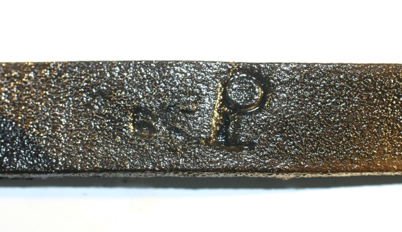 Horizontal strip of dark leather against a white background. Just off-centre to the right of the strip is a stamped mark resembling a letter P.