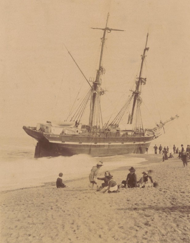 Historic sepia photograph of a two-masted ship to centre of the image, ashore in the surf, with visitors on the beach to foreground and beside the wreck at centre right