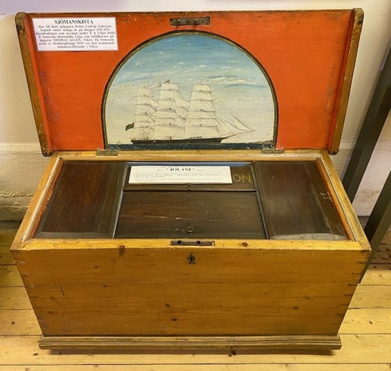 Open wooden sea chest with a portrait of the ship Jolani painted inside the lid in a half-moon set against a painted orange background, and inside the body of the chest three closed dark wood compartments