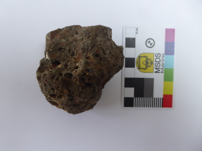 Lump of brown iron slag with cavities in it, with a scale rule to the right showing it to be 8cm high.