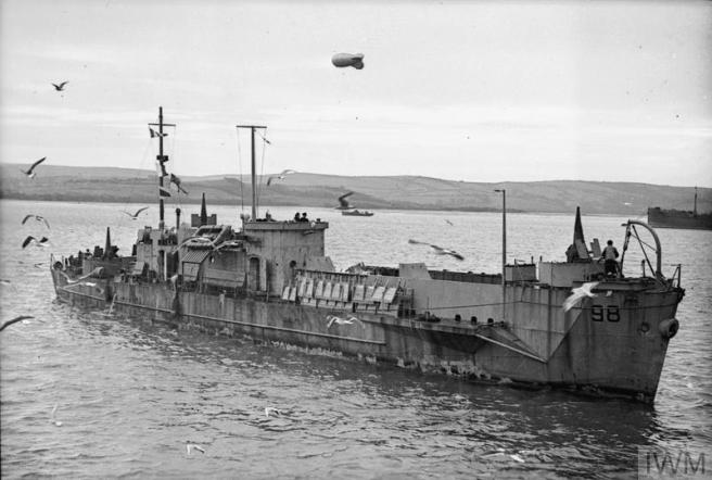 Historic black & white photo of LCI(L) seen in starboard view on the water against a backdrop of hills. Seagulls circle the ship while a barrage balloon flies overhead.