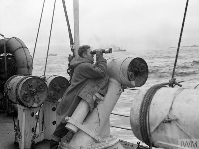 A historic black & white photograph of a man in duffel coat on deck looking out at the convoy with plumes of smoke in the distance, against a swelling sea.