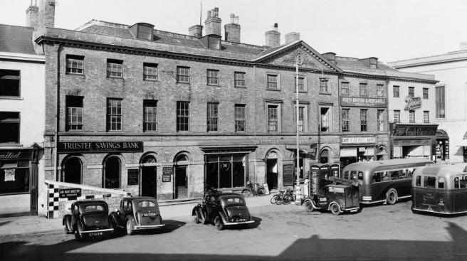 Historic B&W photograph of commercial building surmounted by a triangular pediment. Cars and buses are parked outside and a sign on the building points the way to a public air raid shelter.