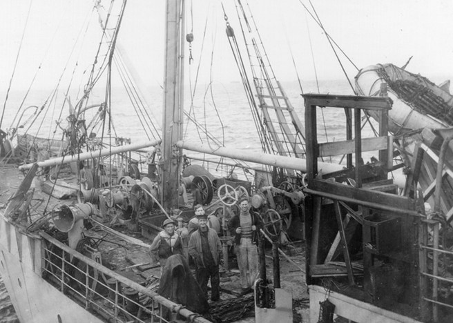 Four crew members standing on the deck of the HMTS Monarch (III) post the attack, surrounded by damaged cable-winding equipment and machinery.