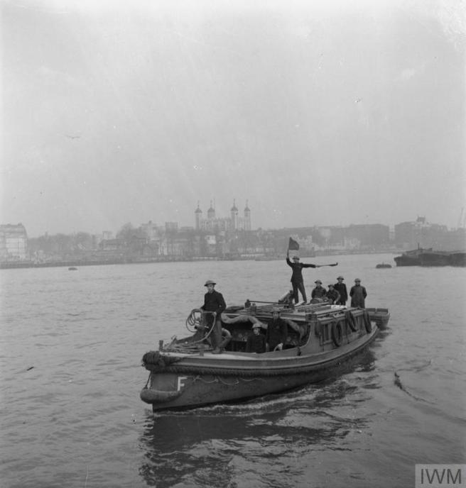 A black and white image showing a group of crew members on a small boat in the river Thames, with some members waving flags. The Tower of London is visible in the background.