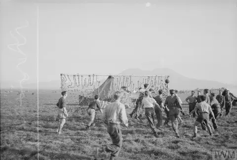 An RAF football match played in Italy between two teams of airmen. Mount Vesuvius is visible in the distance behind the goal made of camouflage netting.