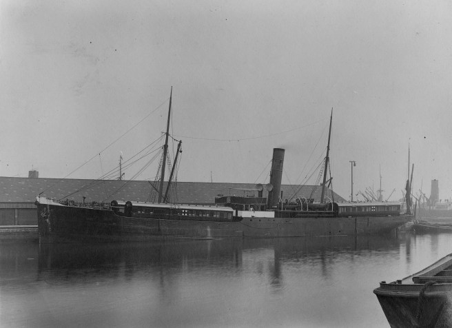 Black and white photo of steamship with single funnel, and four carriages aboard, partly reflected in the water below.