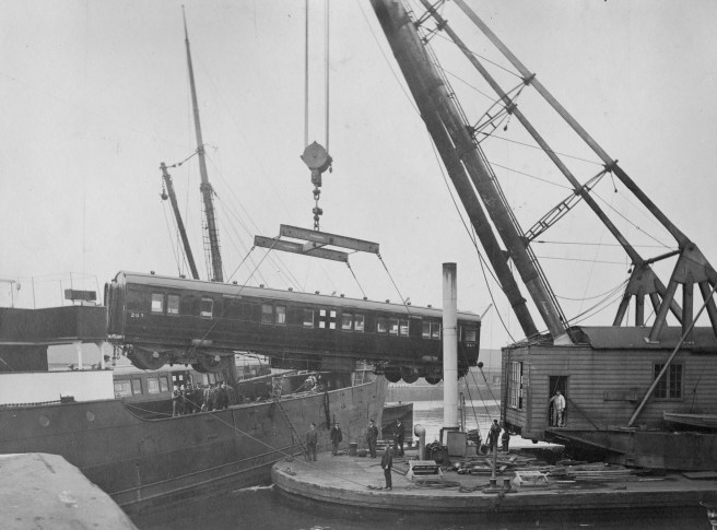 Black and white photograph of crane at right with suspended railway carriage over the deck of a ship.
