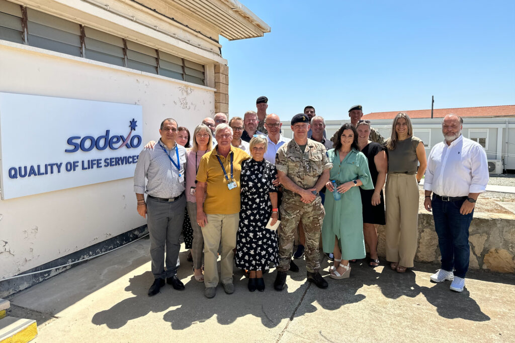 A group of approxiately 18 people stand on a sunny patio next to a Sodexo sign, posing for the camera. Most are in civilian clothes but four are in military uniform.