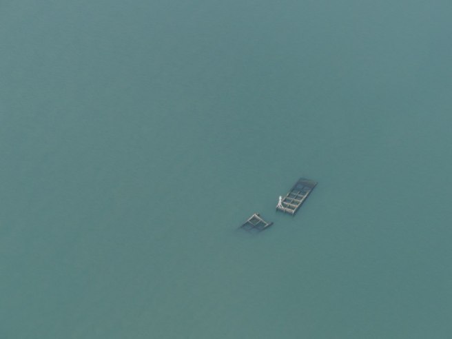 Modern colour photograph: aerial view of two sunken caissons just visible to centre right, emerging from a large expanse of murky grey-green water. A small beacon lies atop one, marking them out as a navigational hazard.