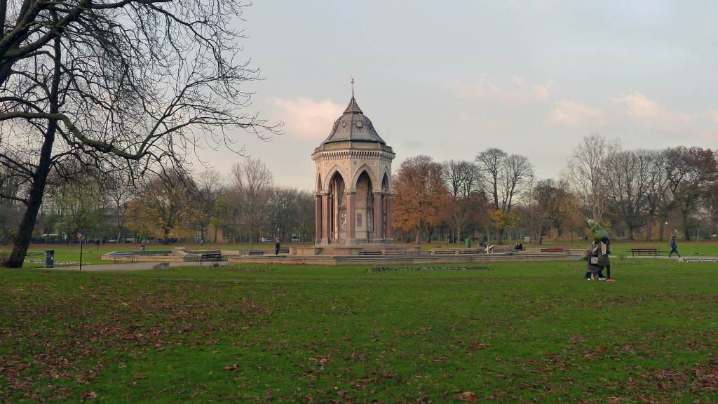 Very tall octagonal canopy with Gothic arches, in a mixture of coloured stones, standing on a stepped base and set in grassy parkland with bare trees in the distance and fallen leaves partially covering the grassy foreground.