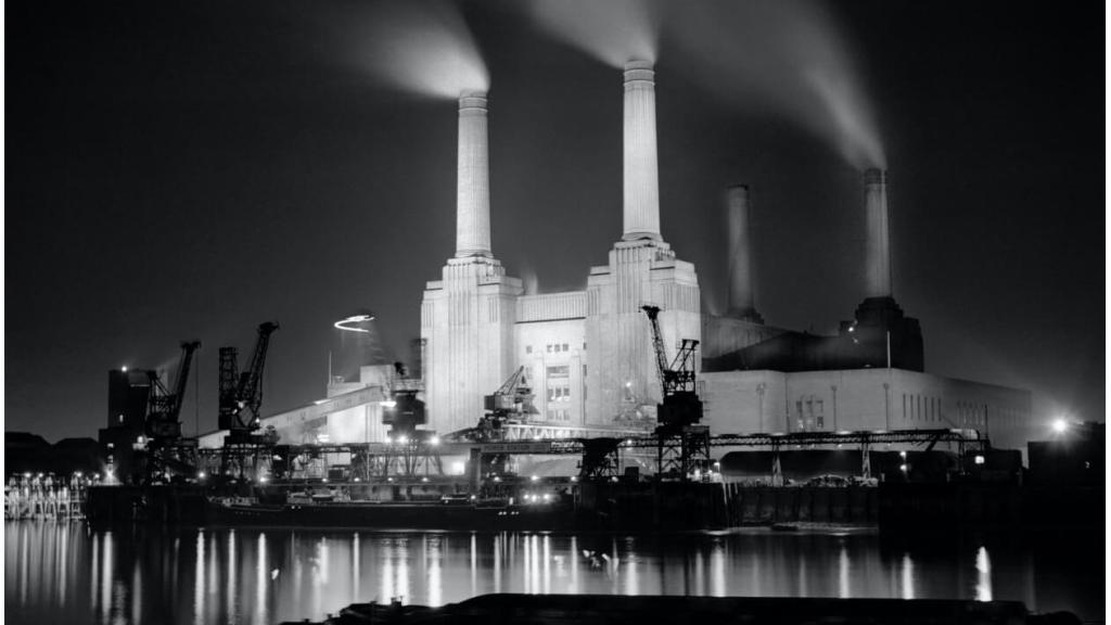 Battersea Power Station at night, viewed across the water of the River Thames, with 3 chimneys belching steam into the black sky.