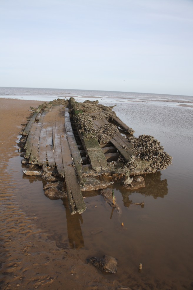 Collapsed hull of wreck still retaining its 'boat shape', half in water to the right, on an extensive beach under a blue sky.