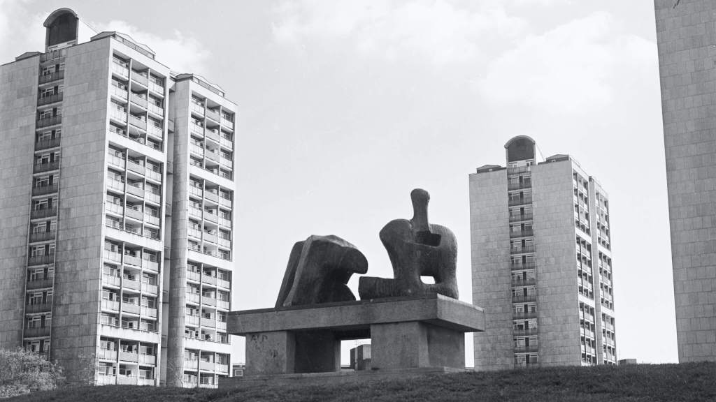 Black and white photo of a Henry Moore sculpture on a concrete plinth in the foreground and 3 tower blocks in the middle distance.