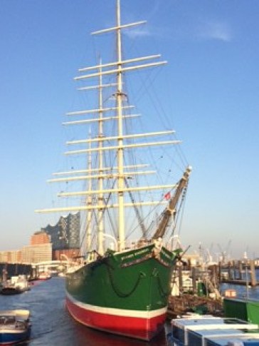 Bow and masts of a tall ship painted green, with a white band and red keel, in harbour, against a blue sky.