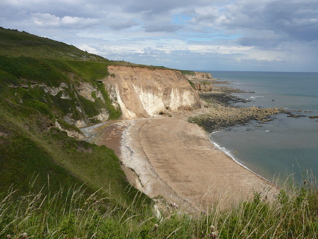 Colour photograph taken from the top of grass-covered cliffs on the left, looking down to an empty beach with bare, vertical cliffs rising from it at centre; to the right rocks and sea, all seen under a cloudy sky.