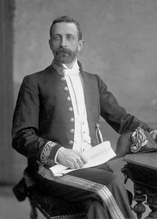 Historic B&W photo of seated man in dark moustache and beard, wearing a formal dress uniform with gold braid to his cuffs and trousers, holding a scroll in his right hand and a gold braided bicorn hat in his left.