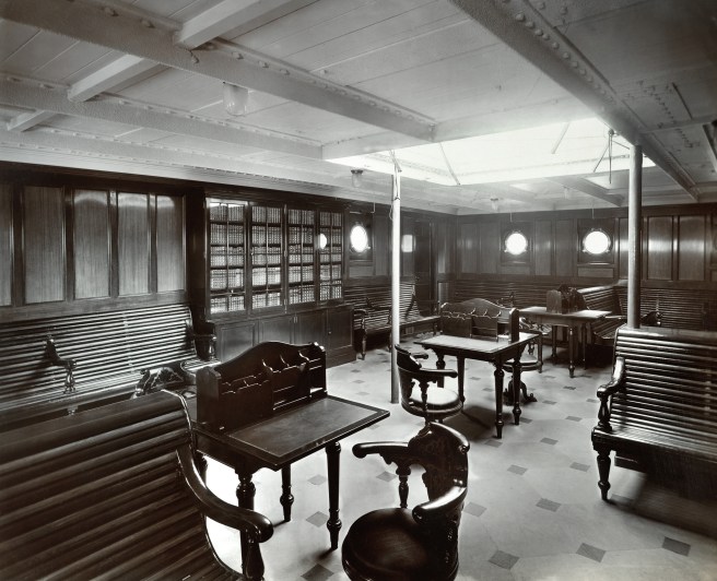 Historic black & white photo of library on board ship, with heavy wooden chairs and desks and lit from above by a skylight cut into the deck above.