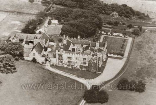 Historic B&W aerial photo of building complex with main courtyard house in centre, set in an open landscape with clumps of trees.