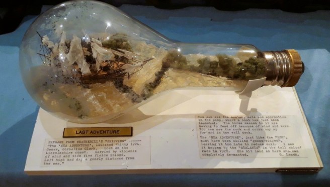 Modern colour photo of a shipwreck in a lightbulb-shaped bottle with a museum label entitled "Last Adventure"