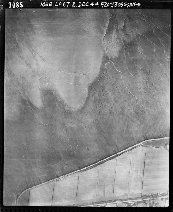 Historic black and white aerial photograph, showing an expanse of marshland at low tide criss-crossed by creek to centre and top of image, with fields of cultivated land to bottom of image.