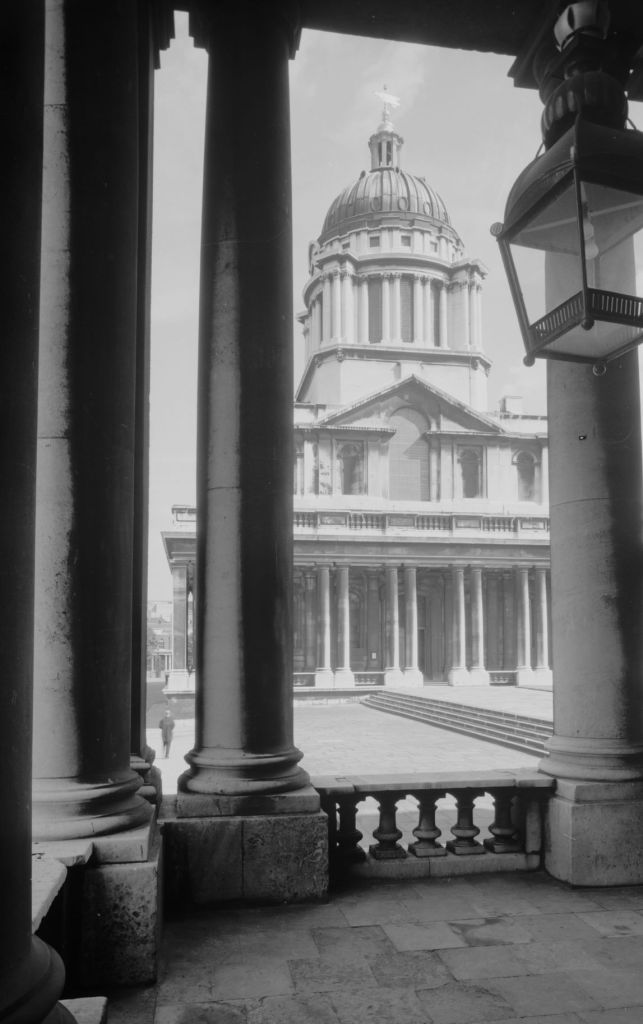 Black and white photograph of colonnaded building with a cupola seen through the columns of a building opposite, and a lamp at top right corner.