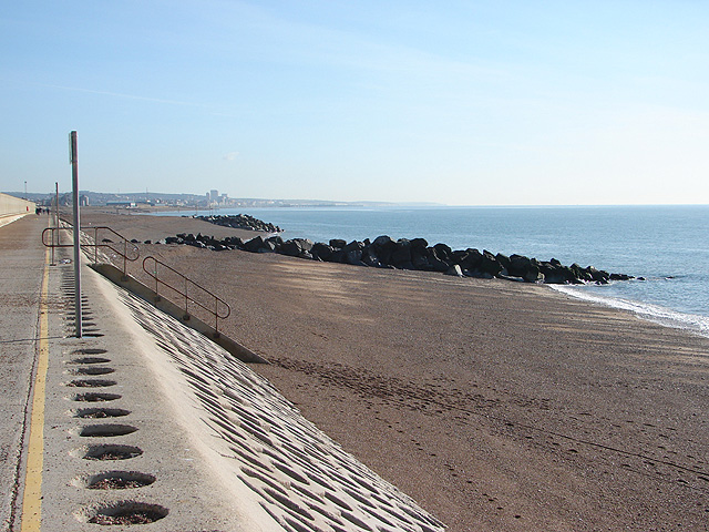 Modern colour photograph on a sunny day taken from a path at the top of the beach (left of image), looking down towards the beach running to the east, (centre of image) broken up by boulder breakwaters at intervals. To the right is a calm blue sea. 