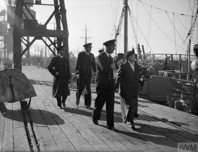 Historic black & white photograph of a dockside scene. Queen Wilhelmina, in a coat and hat and accompanied by 3 men in navy uniform, clutches a large bouquet of flowers in her arm. She walks along the quay with shipping beside her on the right, and dockside infrastructure, such as cranes, to the left and in the background. 