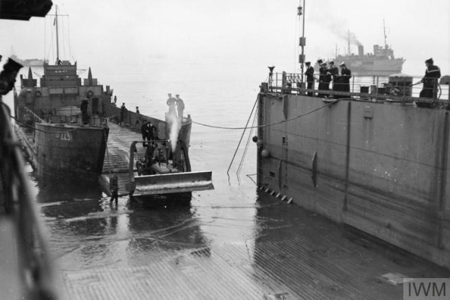 Contemporary black & white photograph of a landing craft tank, ramp down, to left, temporary harbour infrastructure to right, and in the right distance, a steamer waiting offshore.