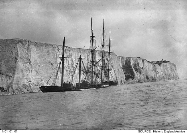 Historic B&W photo of shipwreck against cliffs, seen from seaward, sea filling the lower third of the image.