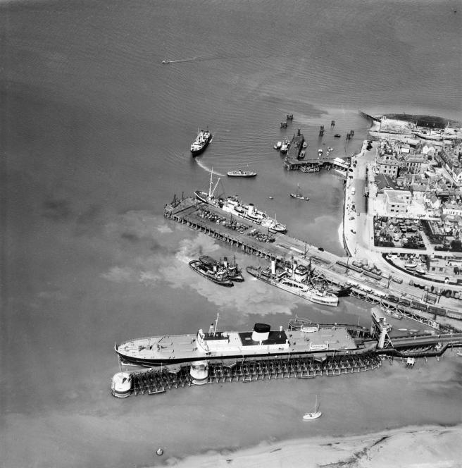 Historic black and white aerial view of Harwich harbour, with the train ferry docked in the foreground, with rails running onto the ship at bottom right. The shoreline is seen at right with other ships berthed or moving about in the harbour.