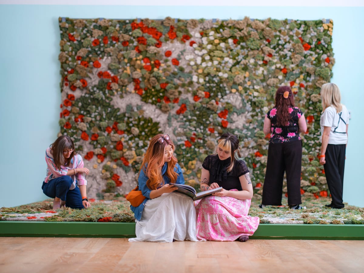 People interacting with an art installation featuring a textured, plant-like wall. Two women sit reading a book, while others observe the display.