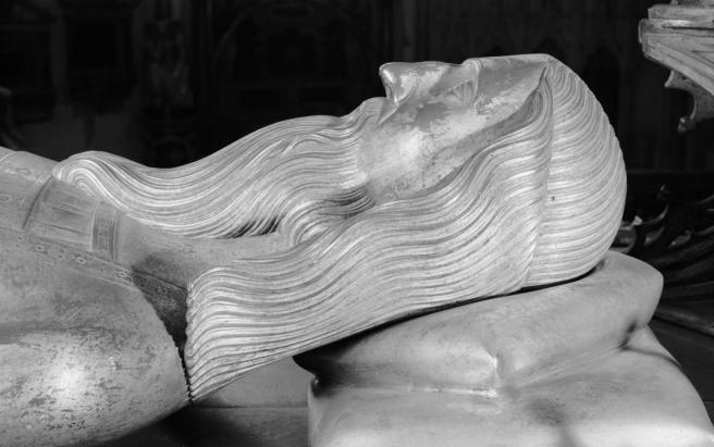Historic mid 20th century photograph in black and white in close-up side view of the effigy head of Edward II, showing his long, wavy hair and beard, a prominent nose, and well-defined eyebrows