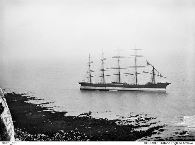 B&W photograph of shipwreck of five-masted vessel seen from cliffs above, the tide receding away from the dark rocky shore to left.